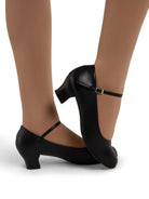 A pair of Capezio Characters shoes in black. Mia Jr 1.5″ black character shoe with PU microfiber upper, 1.5″ sculpted heel, V‑vamp strap, and leather sole styled on foot.